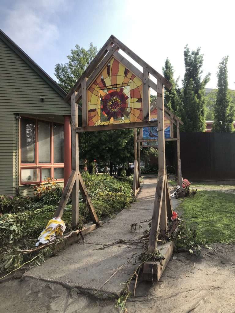 sculpture at Arts Council, a 14-foot wooden frame with stained glass at the top, storm debris around the base