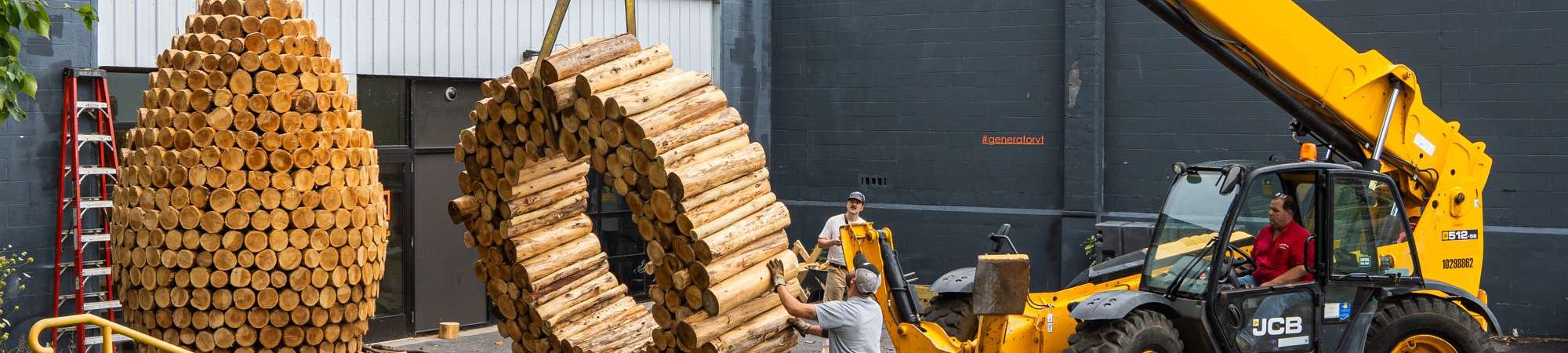 In a parking lot, two people and a yellow long-armed forklift raise a large O made of stacked logs from the ground while a large wooden egg-shaped sculpture sits nearby