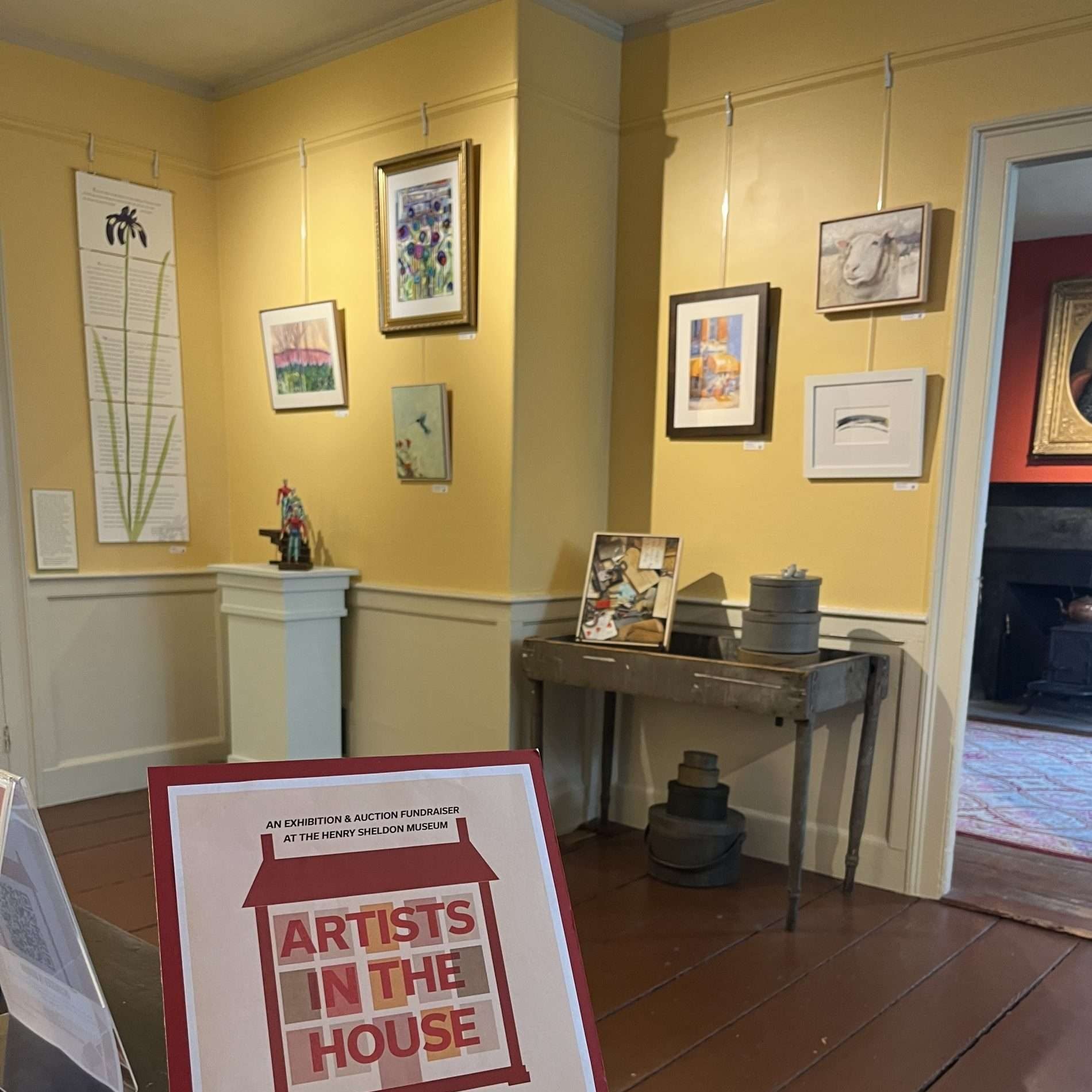 The Henry Sheldon Museum's kitchen room. The walls are yellow and showcase the "Artists in the House Exhibit." In the foreground a poster for the exhibit is displayed.