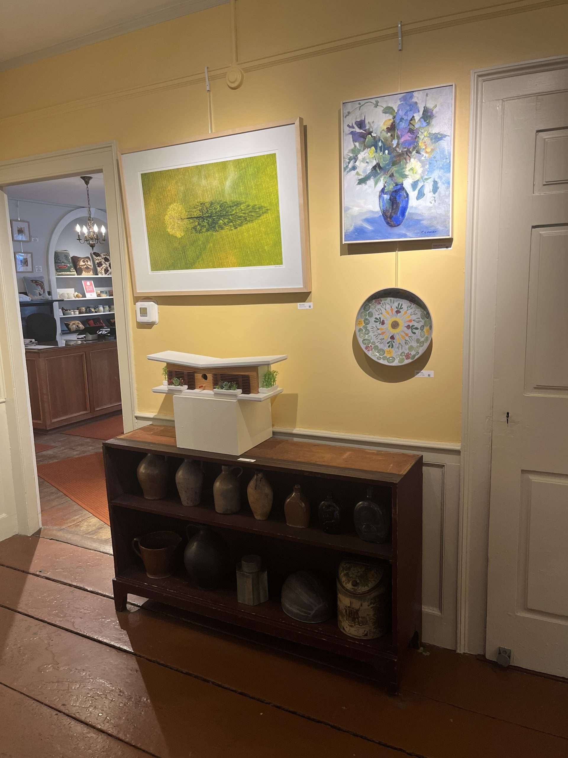 A yellow wall showcasing the art of the "Artists in the House" exhibit. This includes a modern birdhouse, a ceramic bowl, a colorful painting of a flower bouquet, and a photograph of a tree.