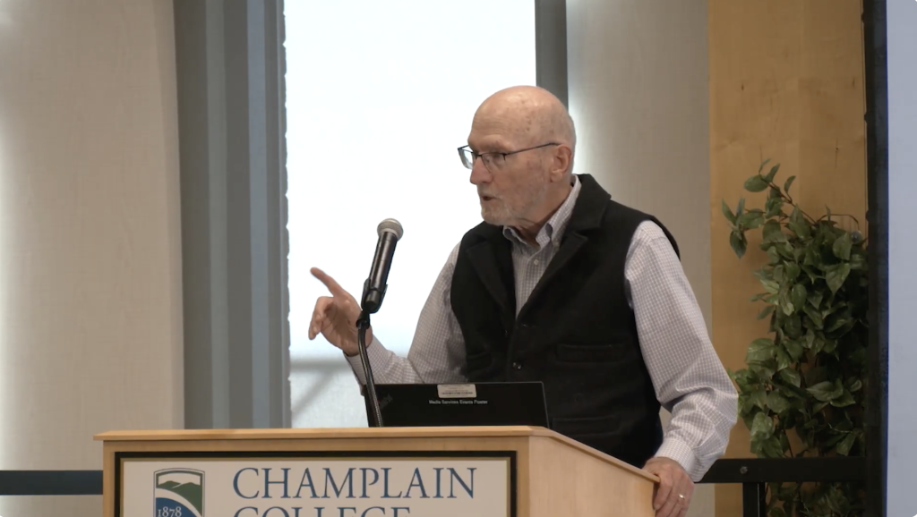 Environmental Author John Elder stands at a podium with a finger raised, looking out into the audience as he reads poetry.